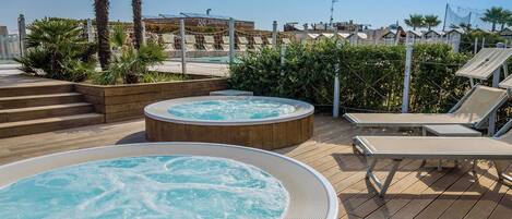 A rooftop pool area with two hot tubs, lounge chairs, and palm trees.