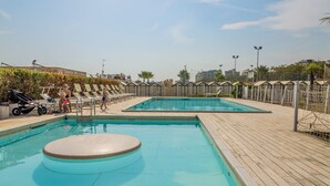 Un área de piscina con dos piscinas, una terraza de madera y una valla con palmeras.
