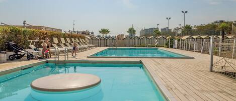 A swimming pool area with two pools, a wooden deck, and a fence with palm trees.