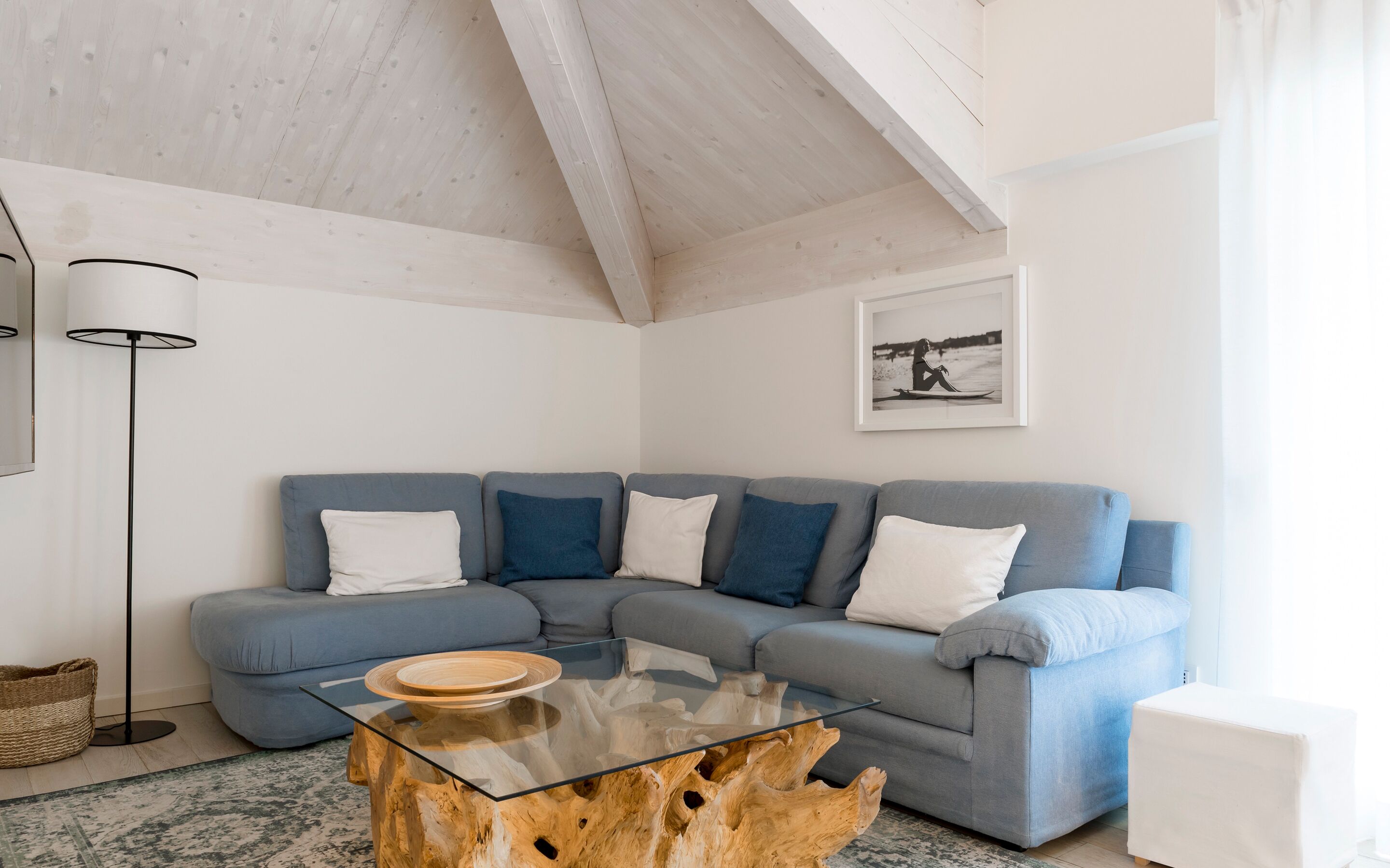 A modern living room with a sectional sofa, a glass coffee table with a wooden base, a floor lamp, and a framed picture on the wall.