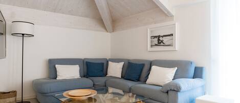 A modern living room with a sectional sofa, a glass coffee table with a wooden base, a floor lamp, and a framed picture on the wall.