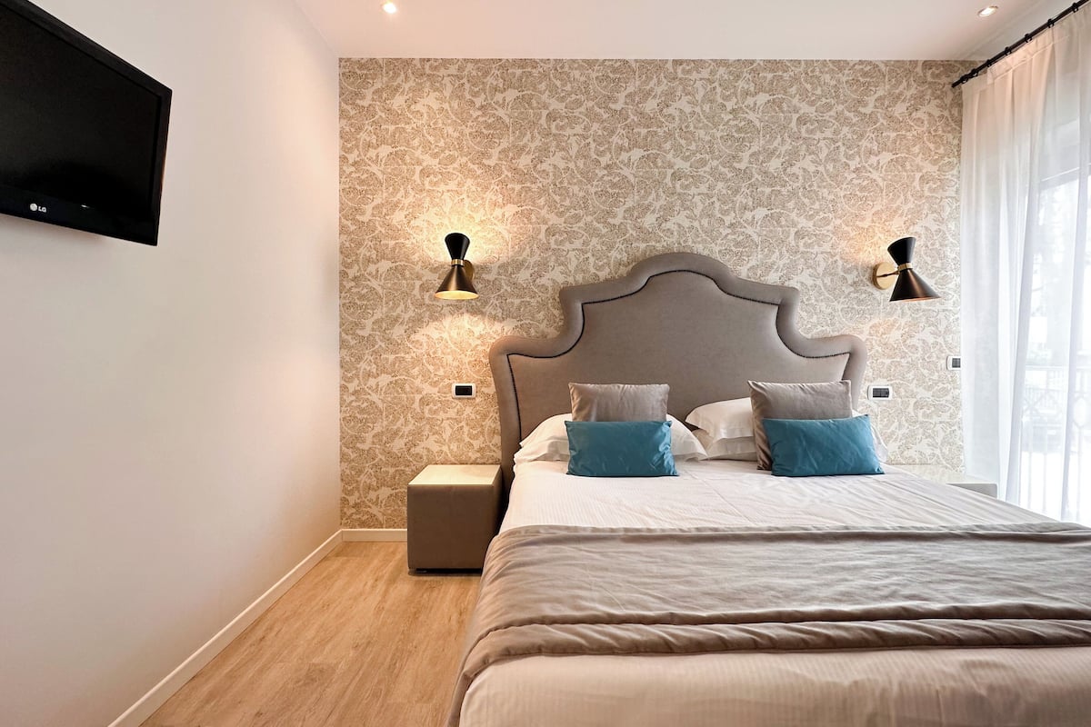 A modern bedroom with a large bed, patterned wallpaper, two wall-mounted lamps, and a TV on the wall.
