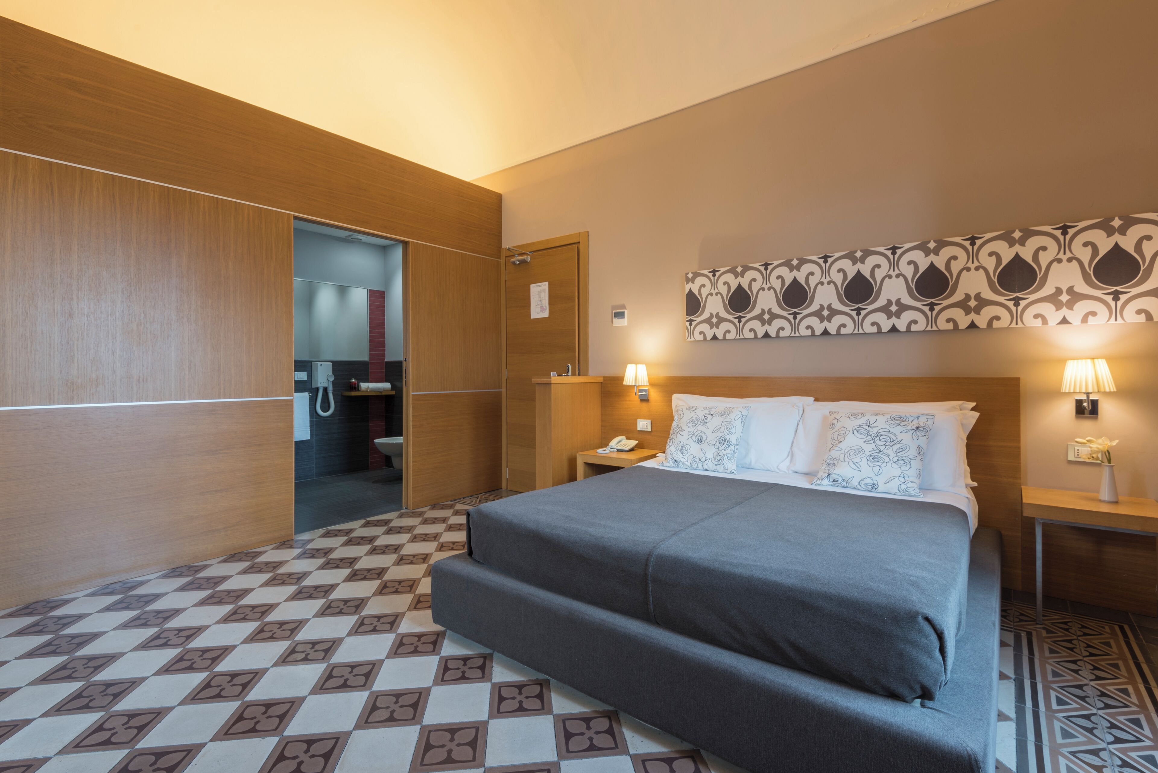 Une chambre d’hôtel avec un lit, une table de chevet, une lampe fixée au mur et un sol à motifs.