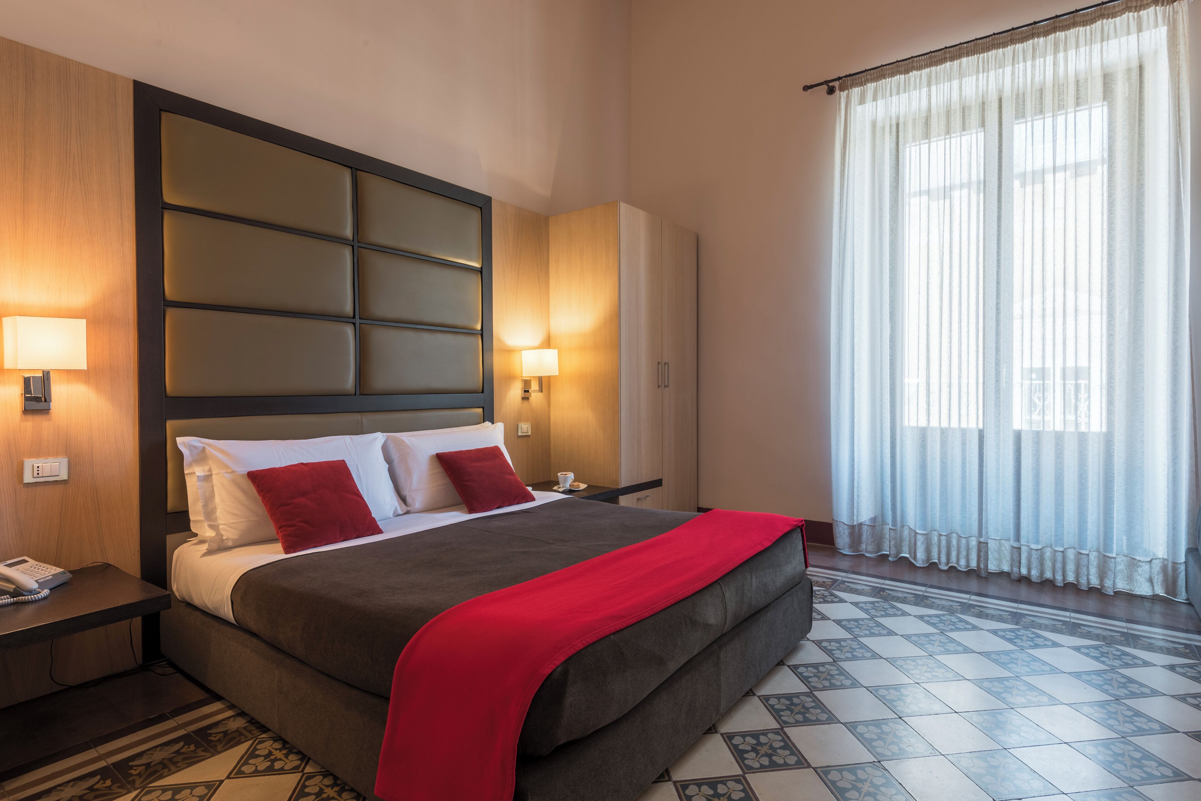 Une chambre d’hôtel avec un grand lit, une table de chevet avec un téléphone, une armoire et une fenêtre avec des rideaux légers.