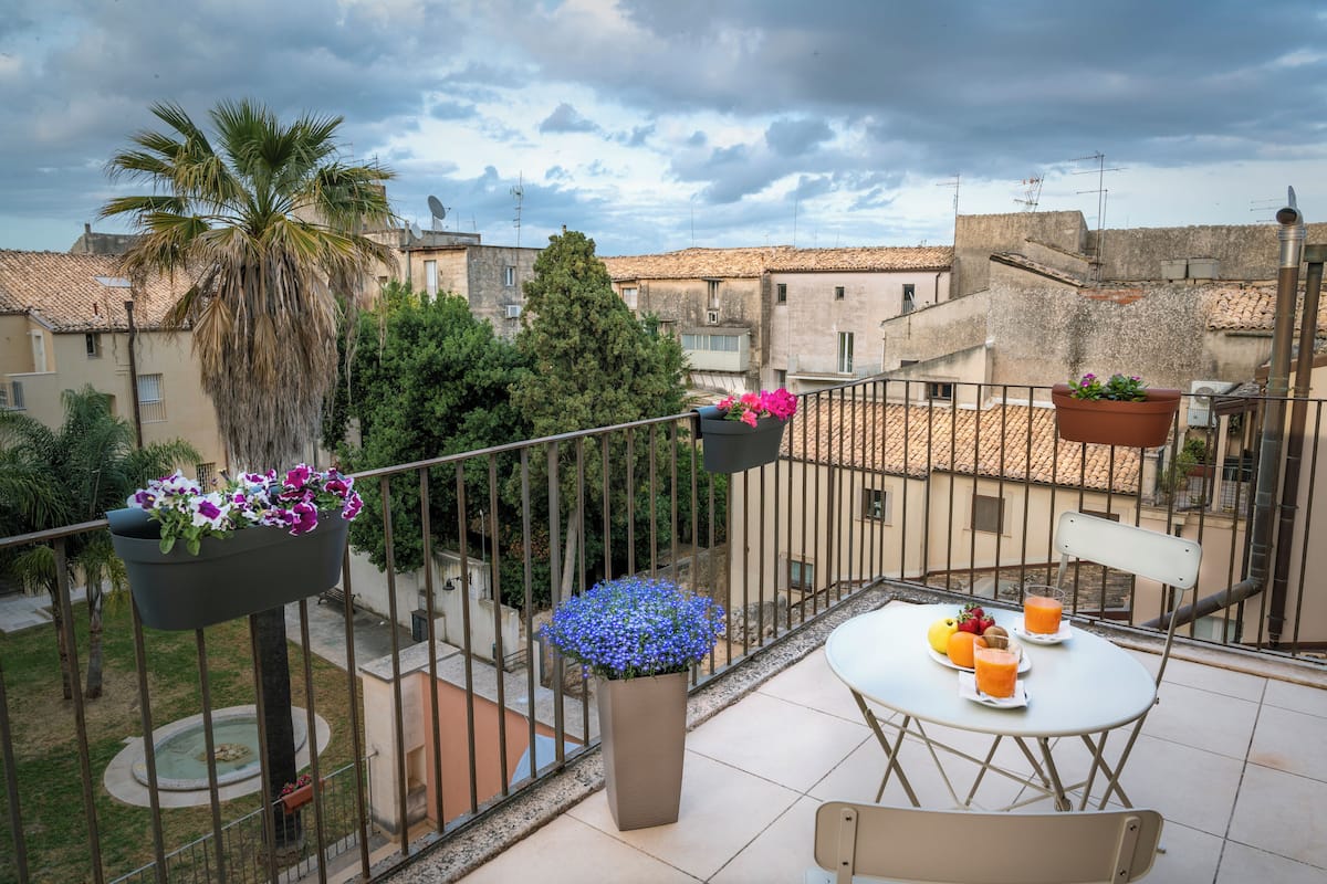 A balcony with a table set for two, potted plants, and a view of the surrounding buildings.