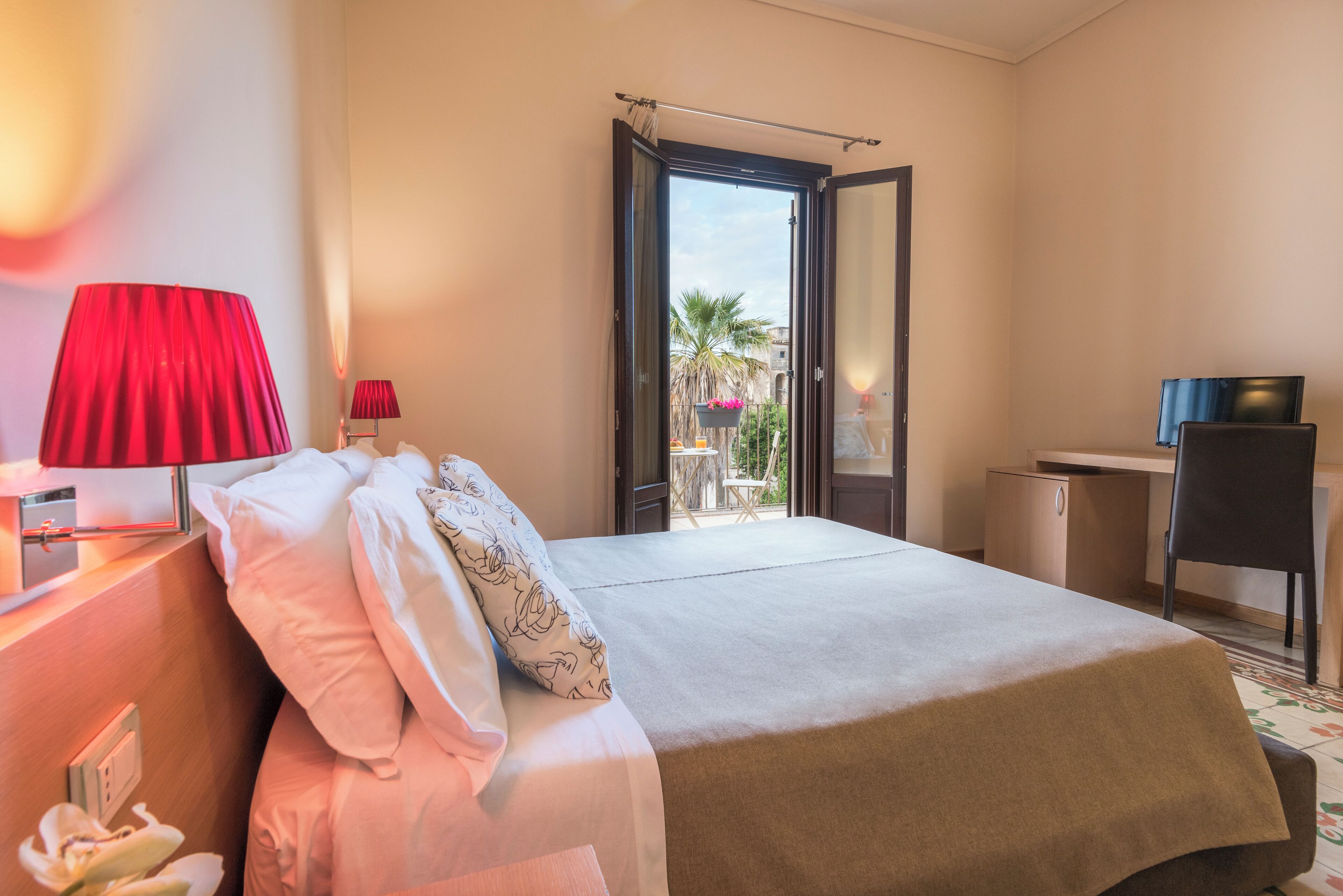 A hotel room with a bed, two bedside lamps, a desk, and an open door leading to a balcony with a view of palm trees and a building.