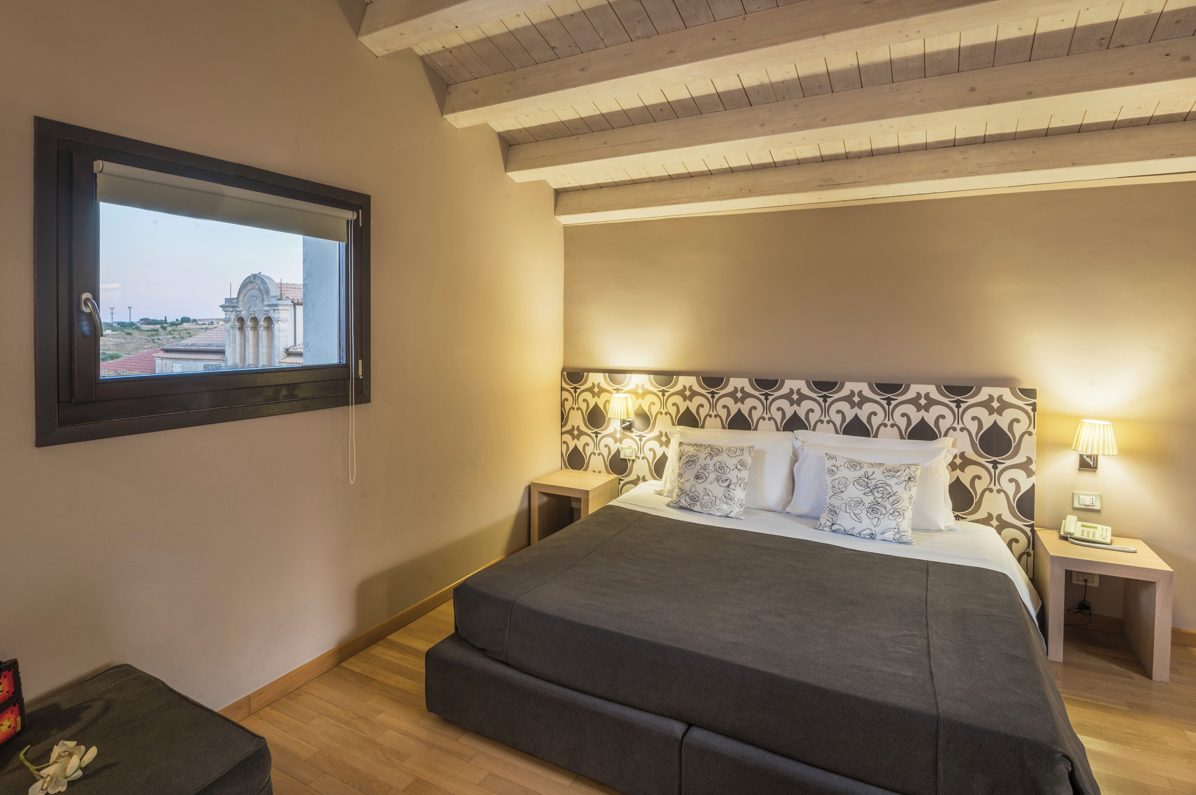 A bedroom with a large bed, a nightstand with a lamp, a small table, and a window with a view of a building.