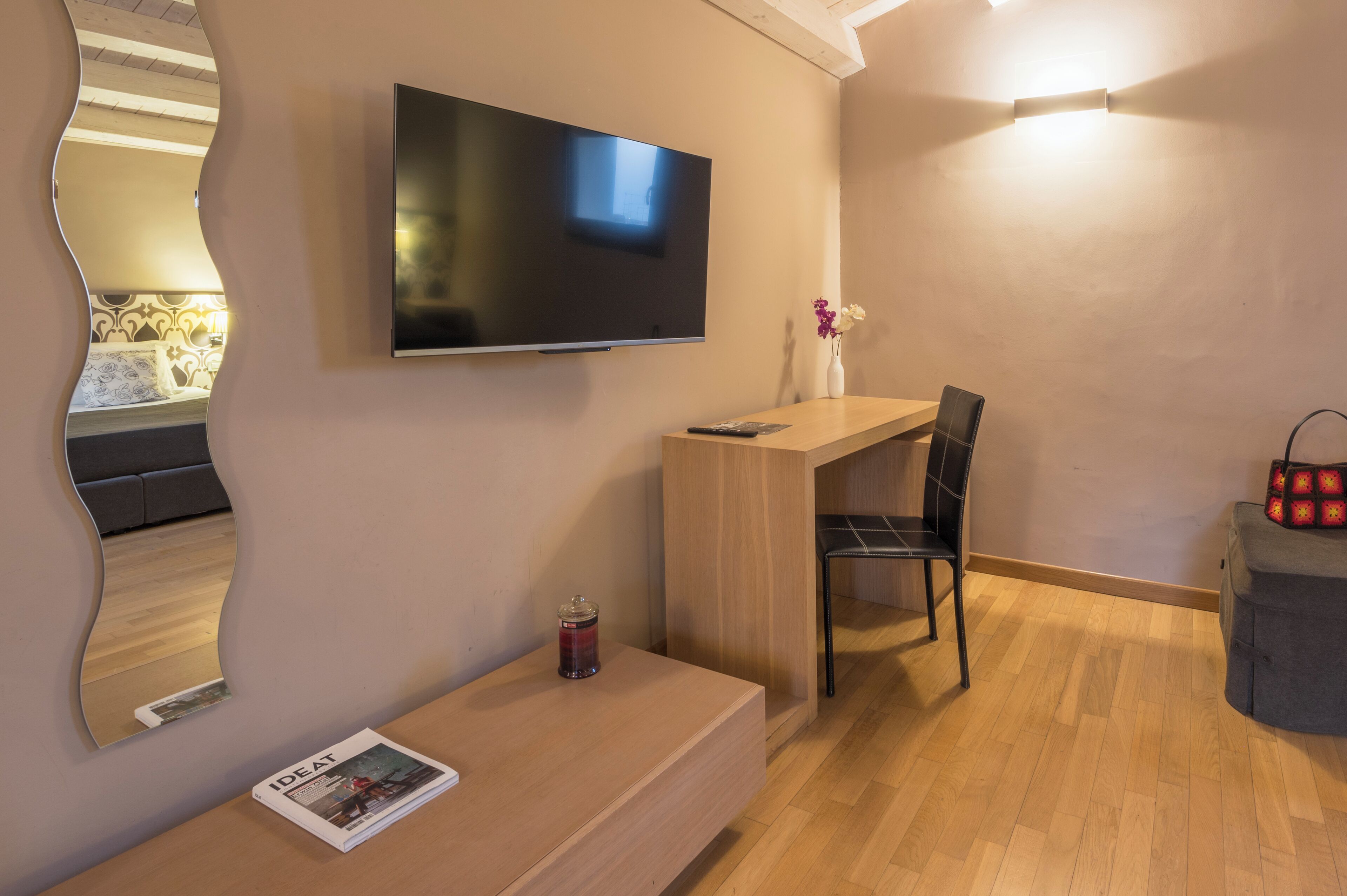 Ein modernes Zimmer mit Flachbildfernseher an der Wand, einem Holztisch, einem schwarzen Stuhl, einem kleinen Tisch mit einer Zeitschrift und einer Vase mit Blumen.