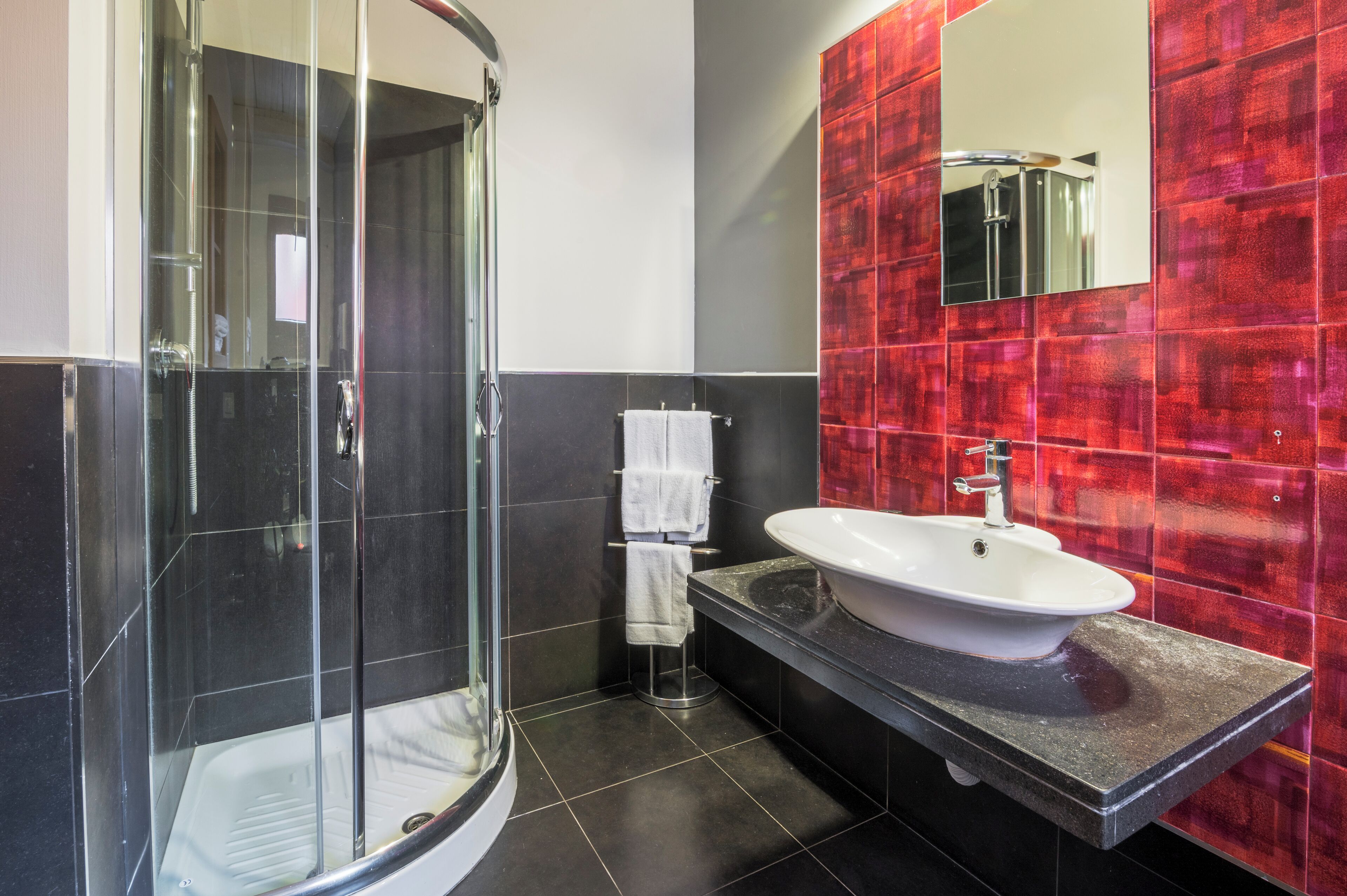 A modern bathroom with a glass shower enclosure, a white sink, and a red-tiled wall.
