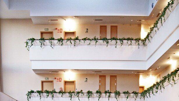 Een ruime lobby met verschillende zithoeken, een hoog plafond met dakramen, en een balkon op de tweede verdieping met beplanting.