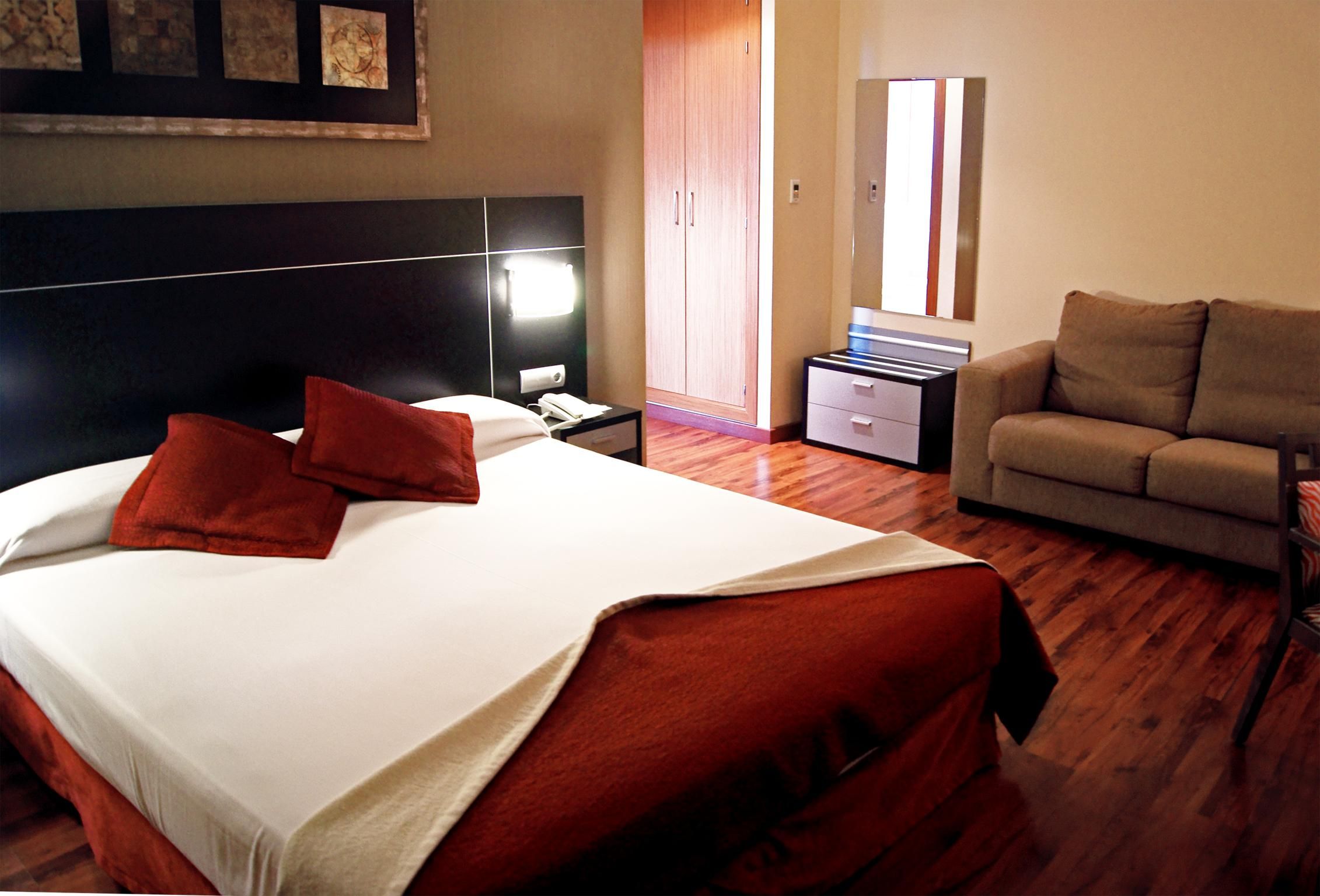 A hotel room with a large bed, a bedside table with a phone, a nightstand, a sofa, and a wardrobe.