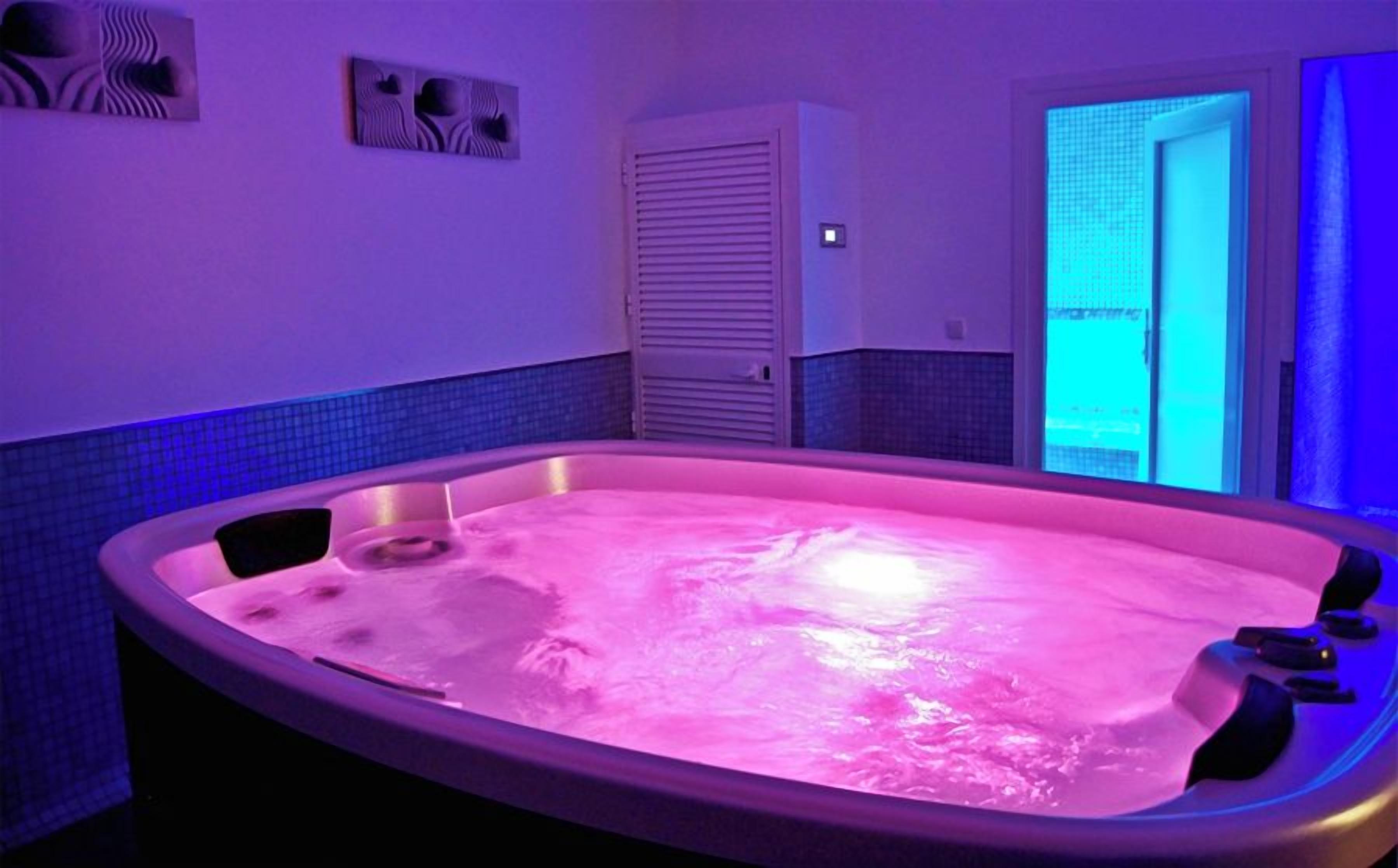 A hot tub with a pinkish hue, surrounded by a tiled wall with a blue accent, and a door leading to an outdoor area.