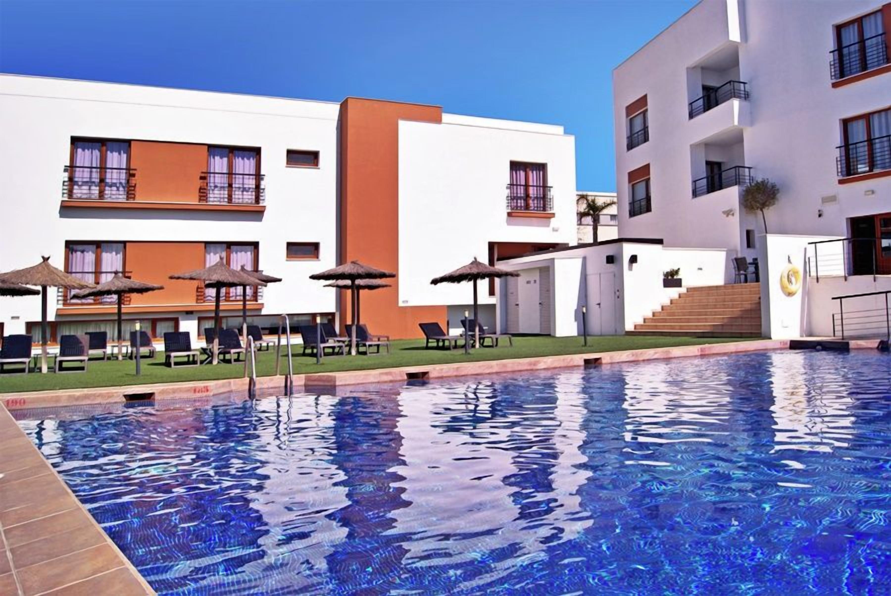 Una piscina con lettini e ombrelloni, circondata da edifici moderni con balconi.