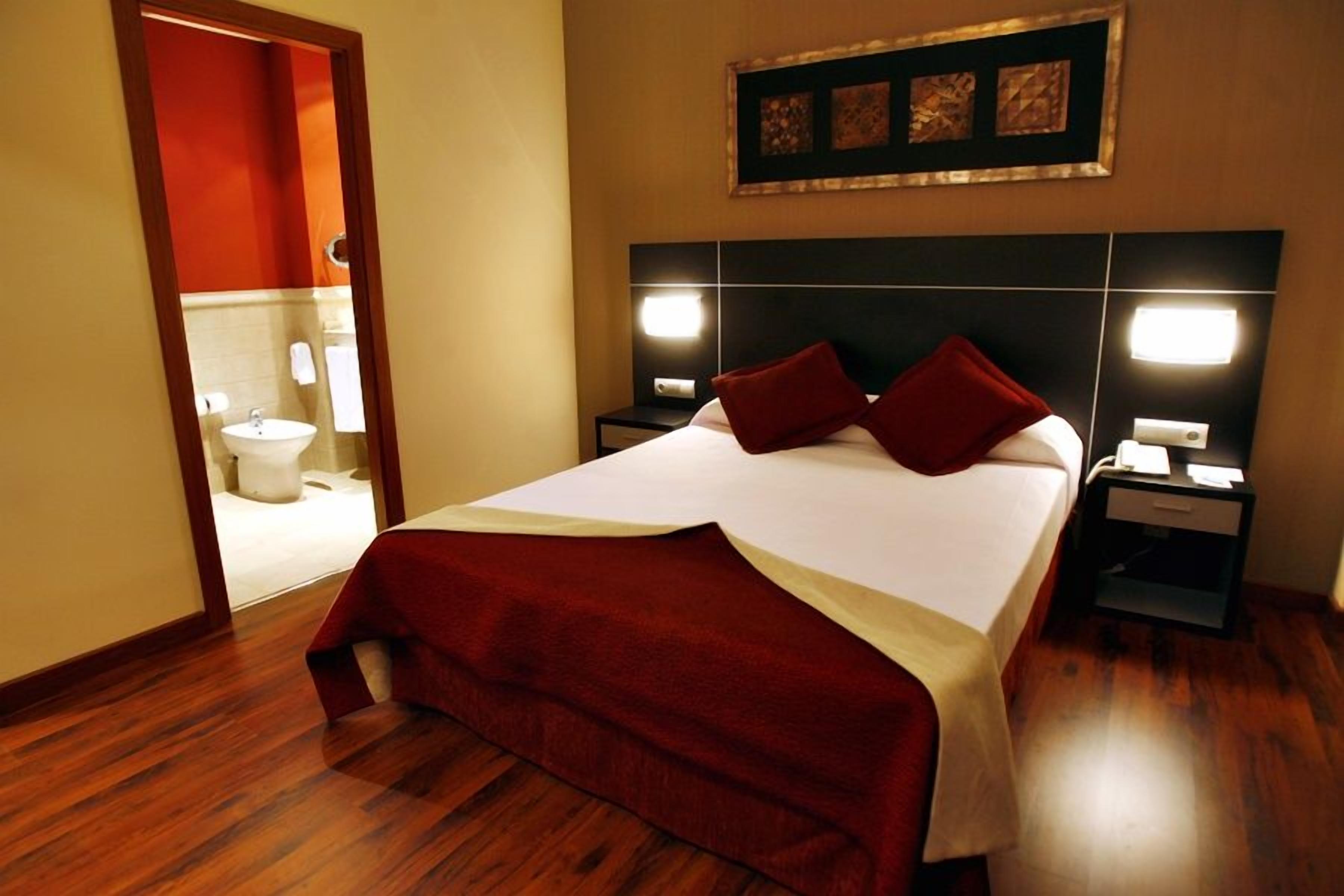 Een hotelkamer met een groot bed, twee nachtkastjes, een hoofdbord met ingebouwde verlichting, een rode plaid en een kunstwerk boven het bed.