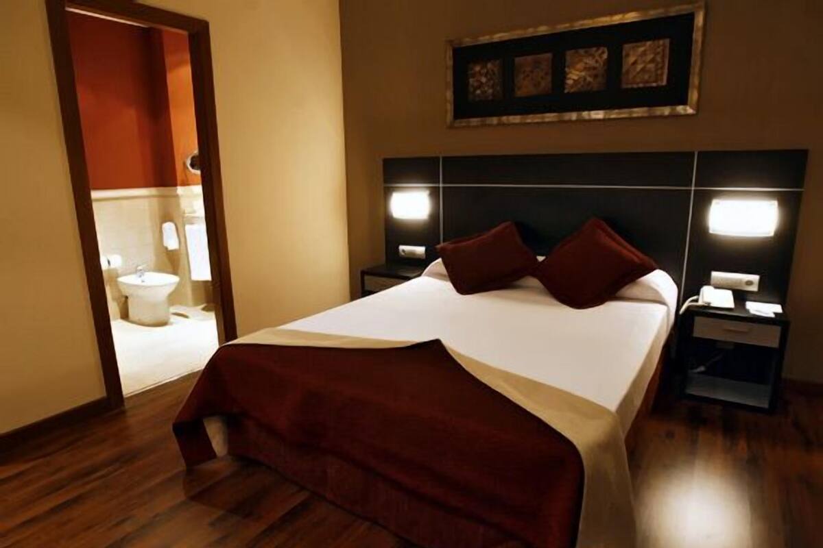 Quarto de hotel com uma cama grande, duas mesas de cabeceira, um criado-mudo, um quadro na parede e um banheiro visível através de uma porta aberta.