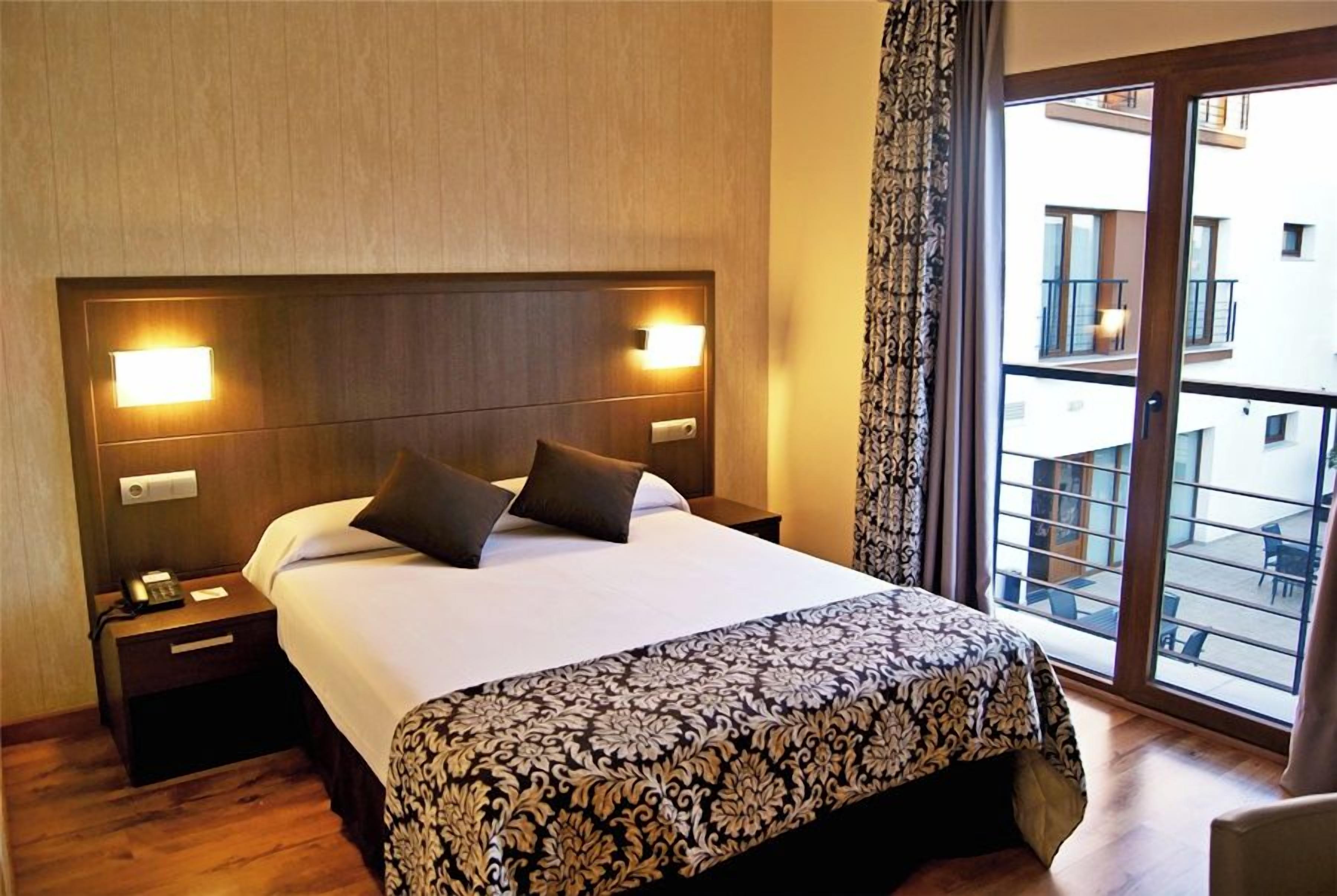 A hotel room with a large bed, a nightstand with a telephone, and a view of a balcony.