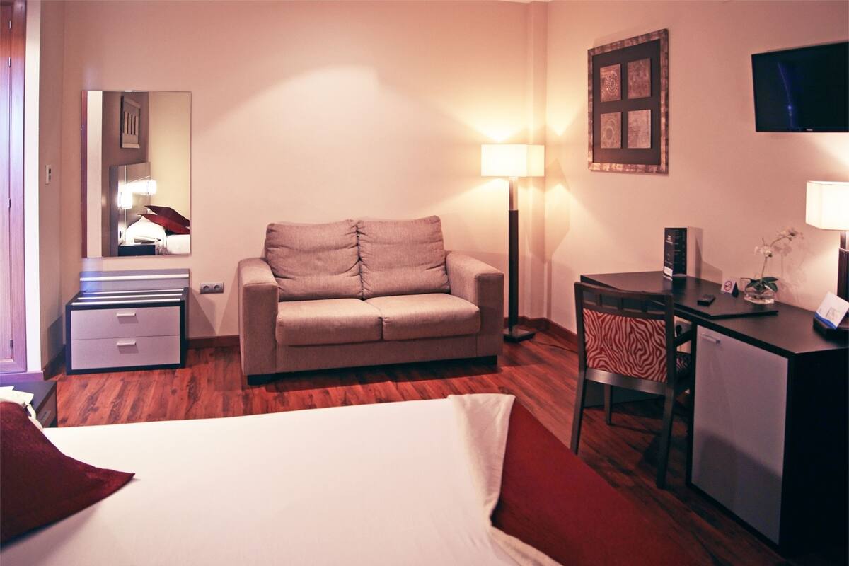 Quarto de hotel com cama, sofá, escrivaninha, cadeira, luminária, televisão e espelho.