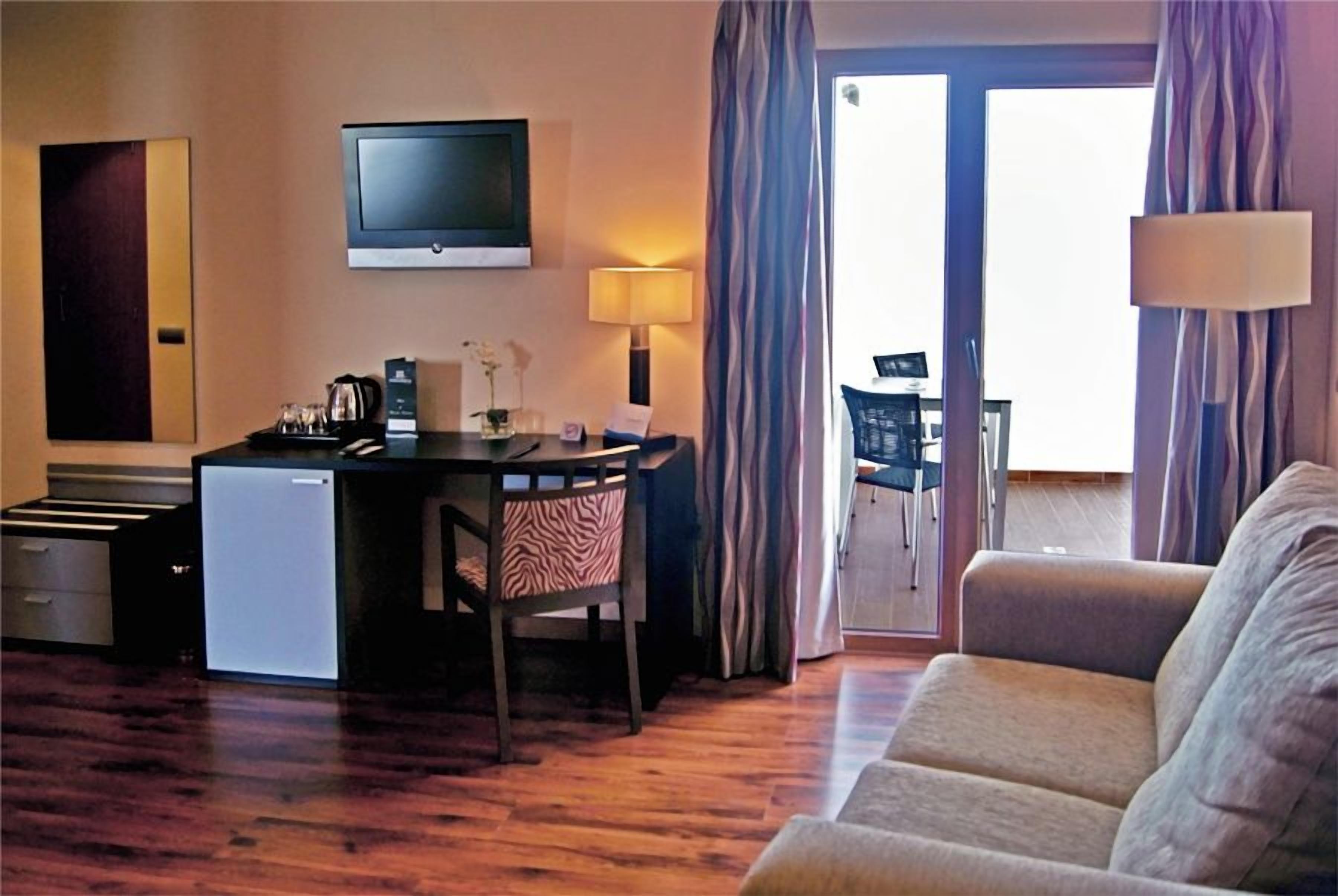 A hotel room with a kitchenette, a flat-screen TV, a sofa, and a view of the outside.