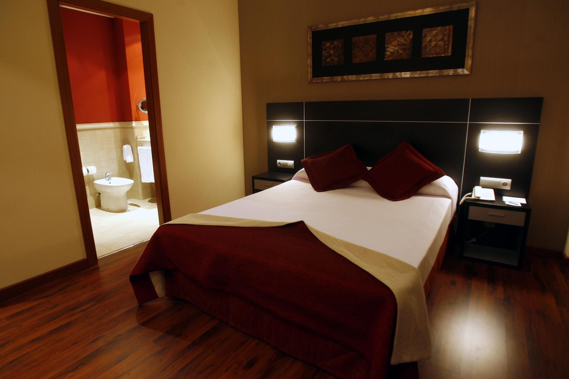 A hotel room with a large bed, two bedside tables, a nightstand, a mirror, and a bathroom visible through an open door.