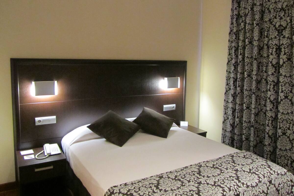 Quarto de hotel com uma cama grande, duas mesas de cabeceira, um telefone e cortina estampada.