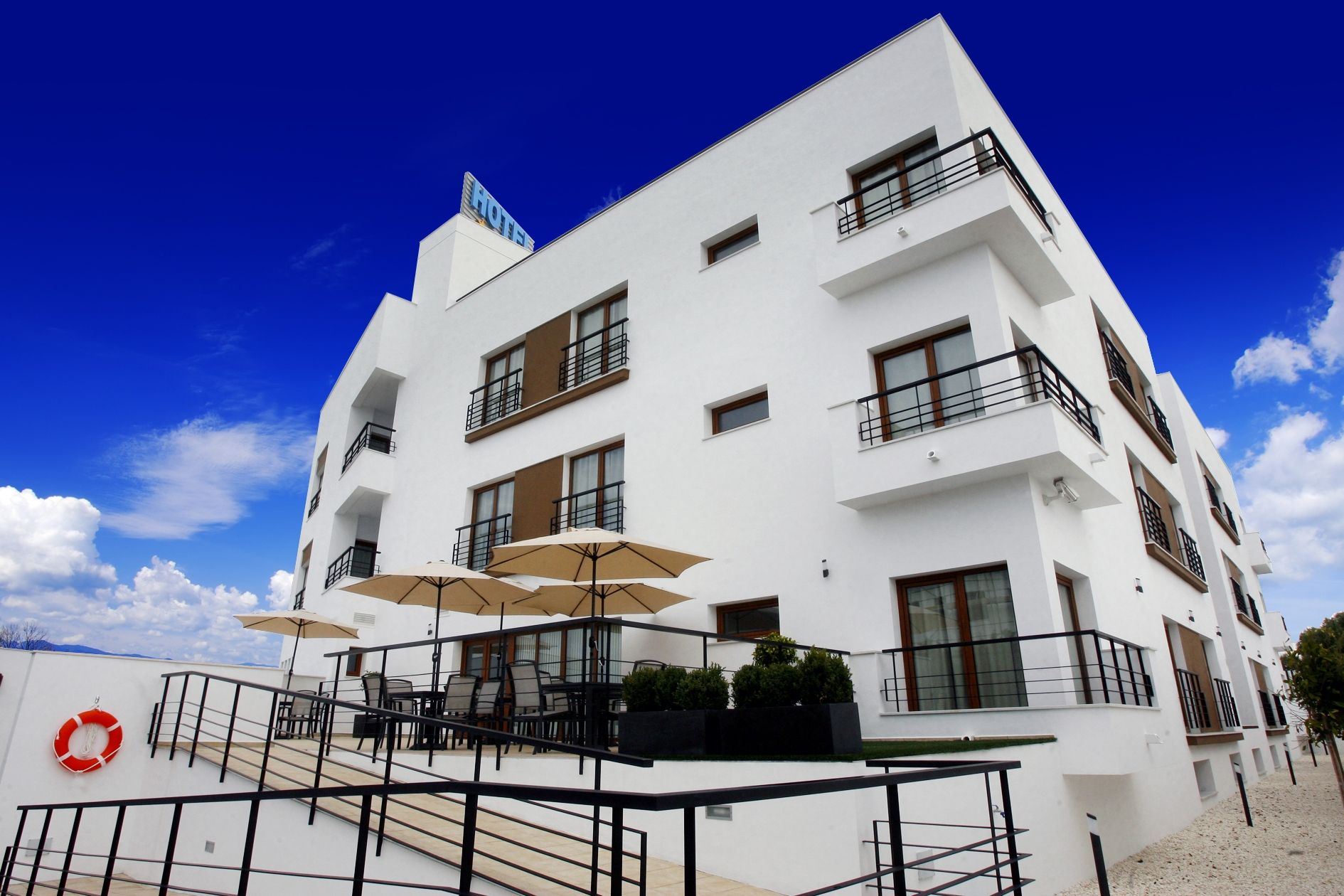 A modern hotel with a white facade, multiple balconies, outdoor seating with umbrellas, and a clear blue sky.