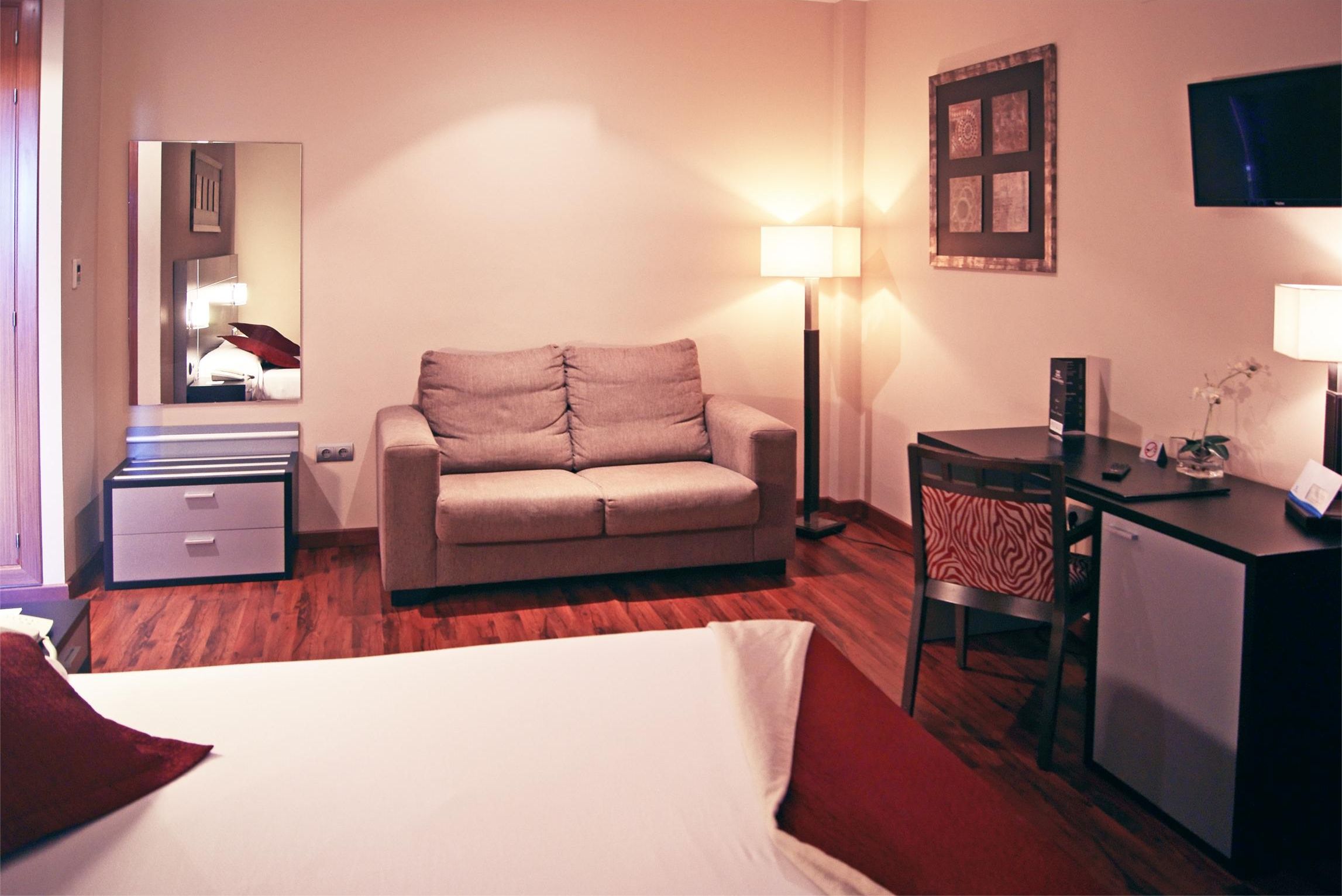 Habitación de hotel con cama, sofá, escritorio, silla, lámpara, televisor y espejo.
