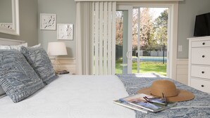 A bedroom with a bed, a nightstand, a lamp, a window with curtains, a hat, and a magazine on the bed.