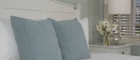 A neatly made bed with two light blue pillows, a white bedside table with a lamp and a vase of flowers, and a window with blinds.
