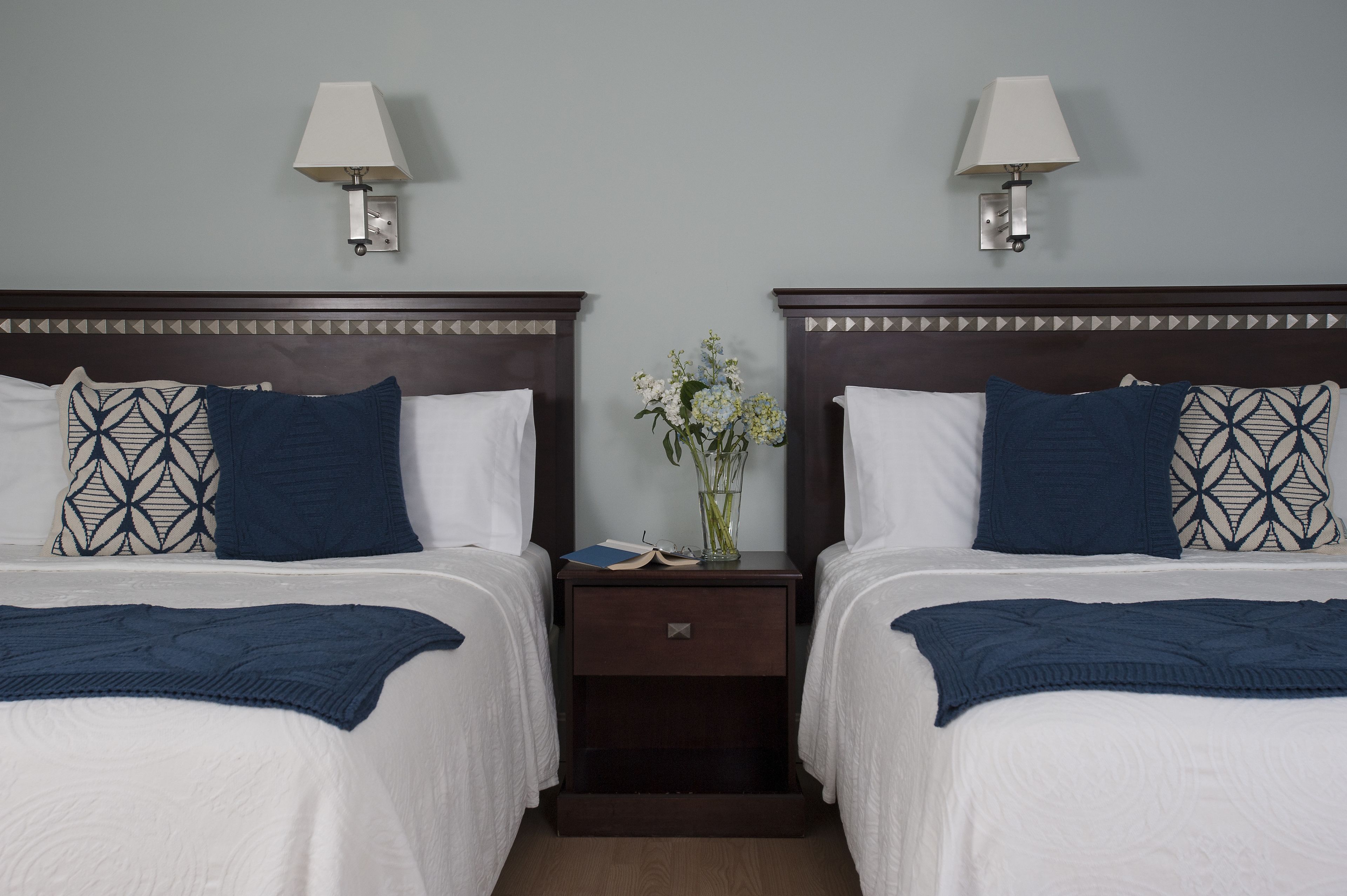 Ein Hotelzimmer mit zwei Betten, jedes mit einem weißen und blauen Kissenmuster, einem dunklen Holz-Kopfstück und einem Nachttisch mit einer Lampe.