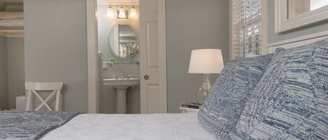 A bedroom with a bed, a nightstand with a lamp, a chair, a mirror, and a bathroom visible through an open door.