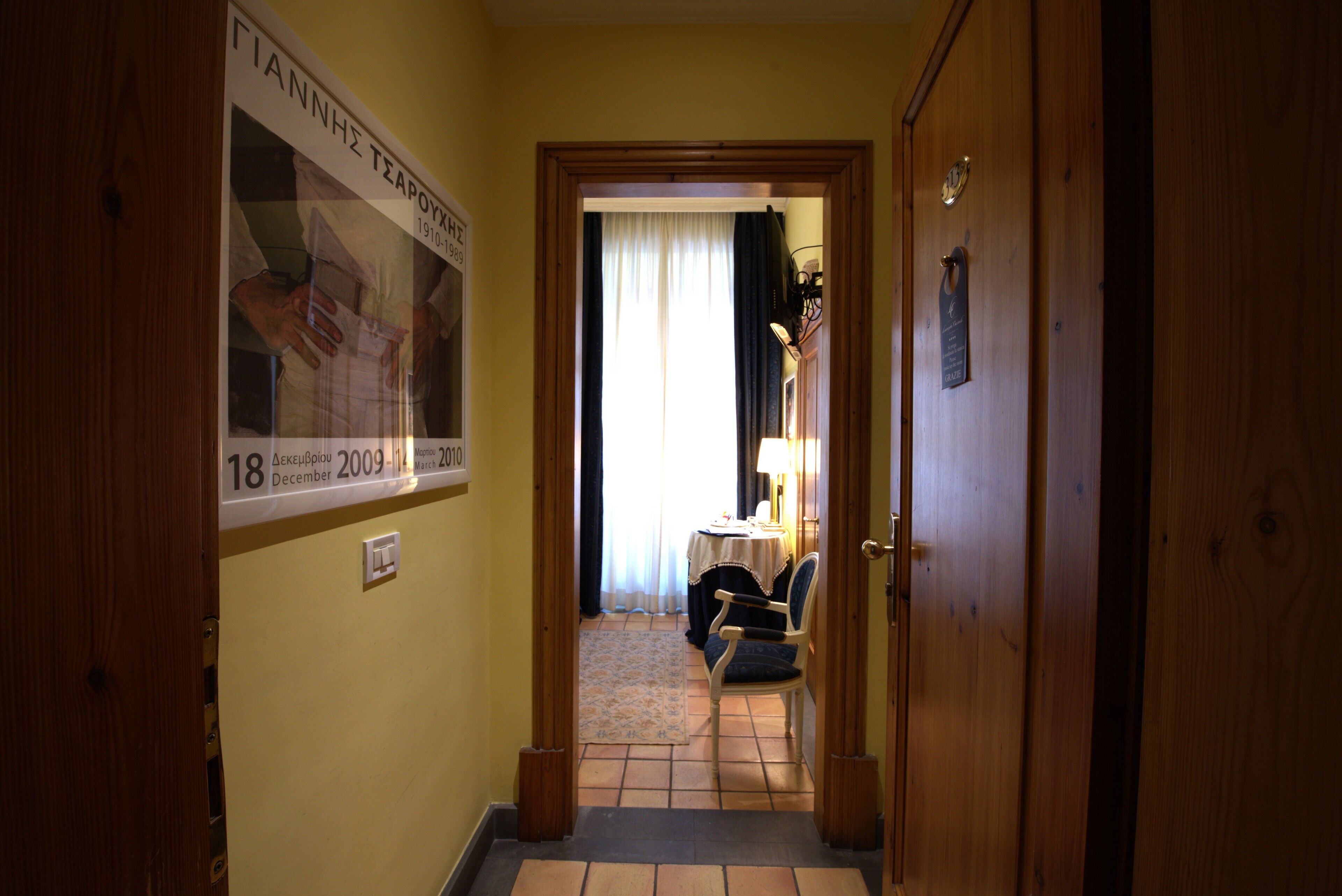 A narrow hallway with a wooden door on the right, a poster on the left, and a room with a table and chair in the background.