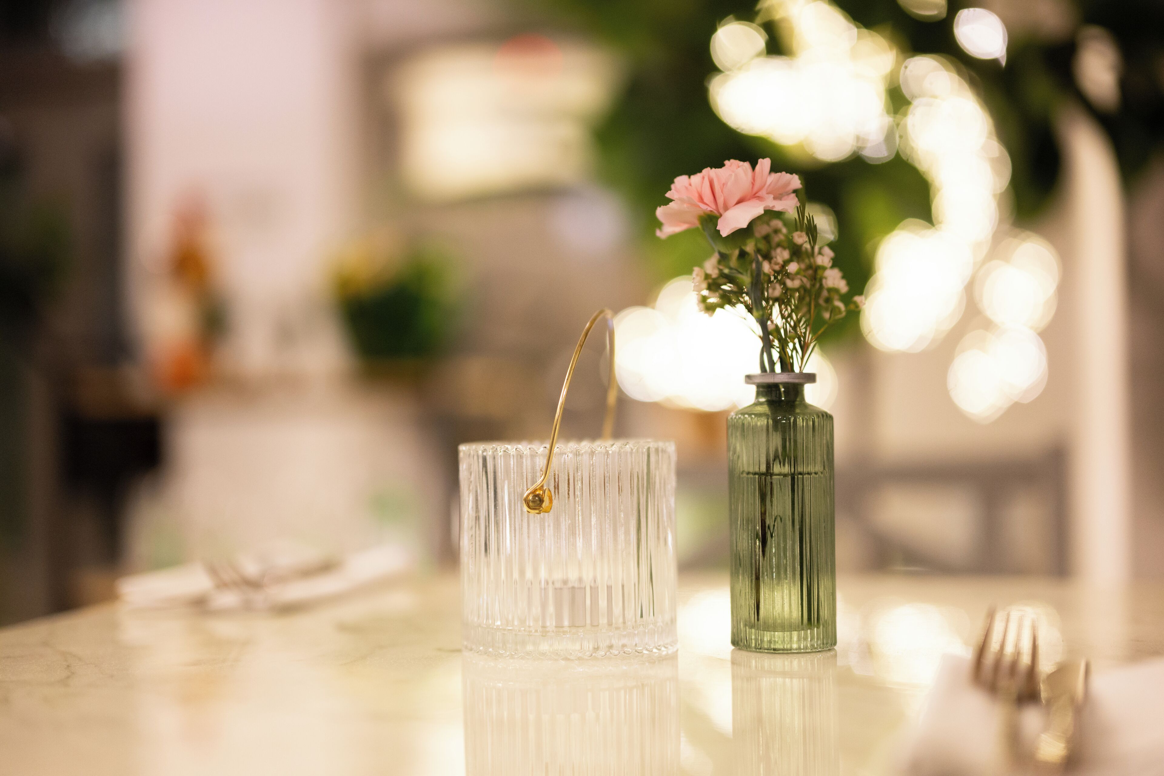 Una mesa con un florero de vidrio transparente con flores rosadas y un florero de vidrio verde con asa dorada.