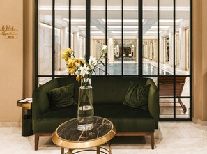 A green sofa with a marble table in front, a vase with flowers, and a view of a pool and lounge area through a glass partition. - Hotel Saski Krakow, Curio Collection by Hilton (Kraków)