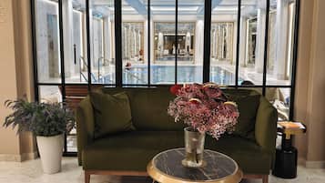 A lobby with a green sofa, a round table with a vase of flowers, and a large window with a view of a pool and a mirrored wall.