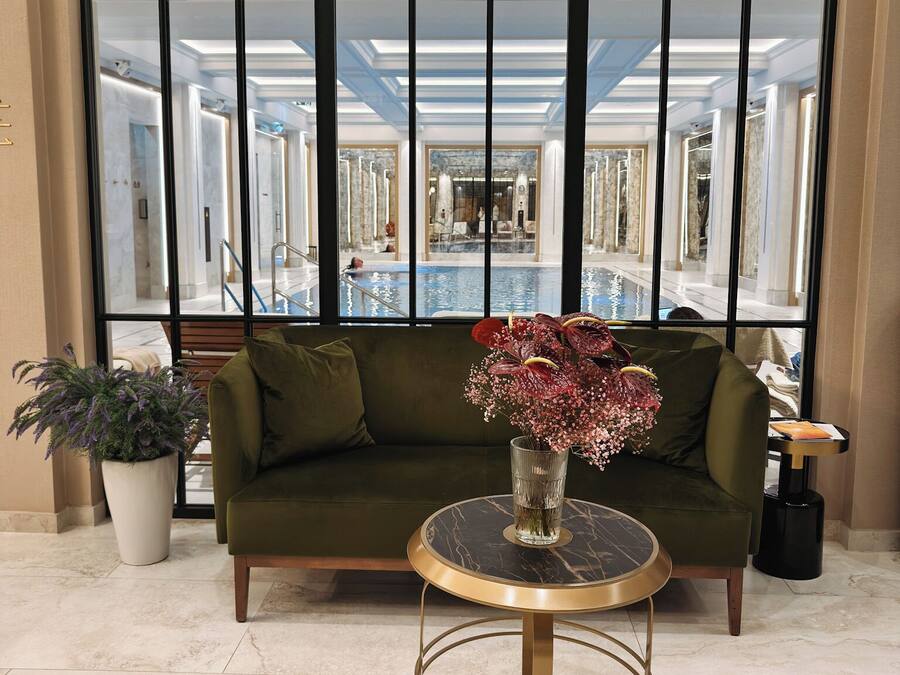 A lobby with a green sofa, a round table with a vase of flowers, and a large window with a view of a pool and a mirrored wall.