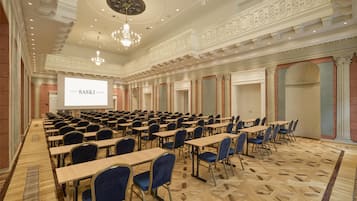 A conference room with rows of tables and chairs, a large screen, and ornate ceiling details.