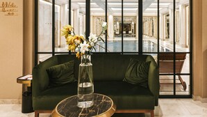 A green sofa with a marble table in front, a vase with flowers, and a view of a pool and lounge area through a glass partition. - Hotel Saski Krakow, Curio Collection by Hilton (Kraków)