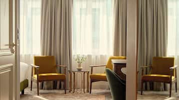 A hotel room with a large window, two yellow armchairs, a green chair, a small table with a vase, and a patterned carpet.