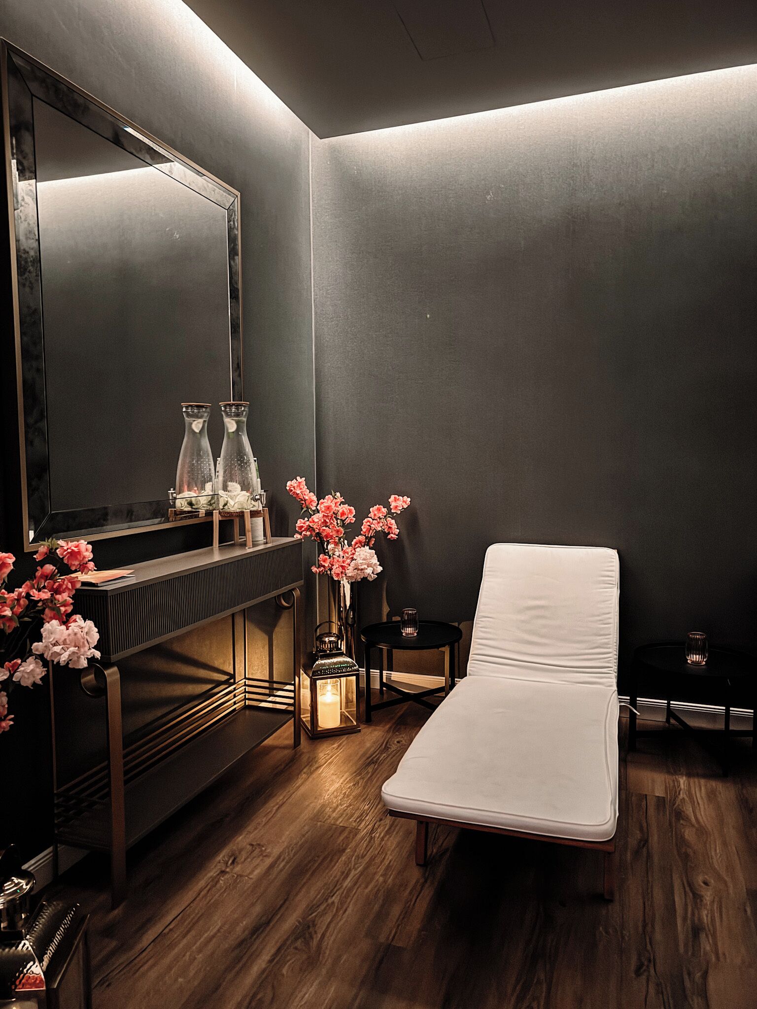 A modern, dimly lit room with a wooden floor, a white massage table, a small side table, a mirror, and two glass bottles on a console table.