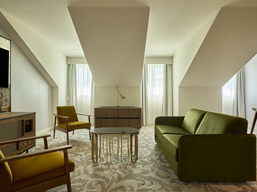 A modern hotel room with a green sofa, yellow armchairs, a wooden cabinet, and a flat-screen TV.
