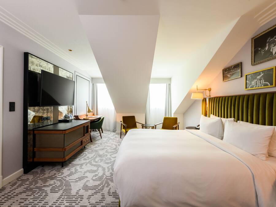 Ein Hotelzimmer mit einem großen Bett, einem Schreibtisch, einem Flachbildfernseher, einem gepolsterten, grünen Kopfteil und einem gemusterten Teppich.