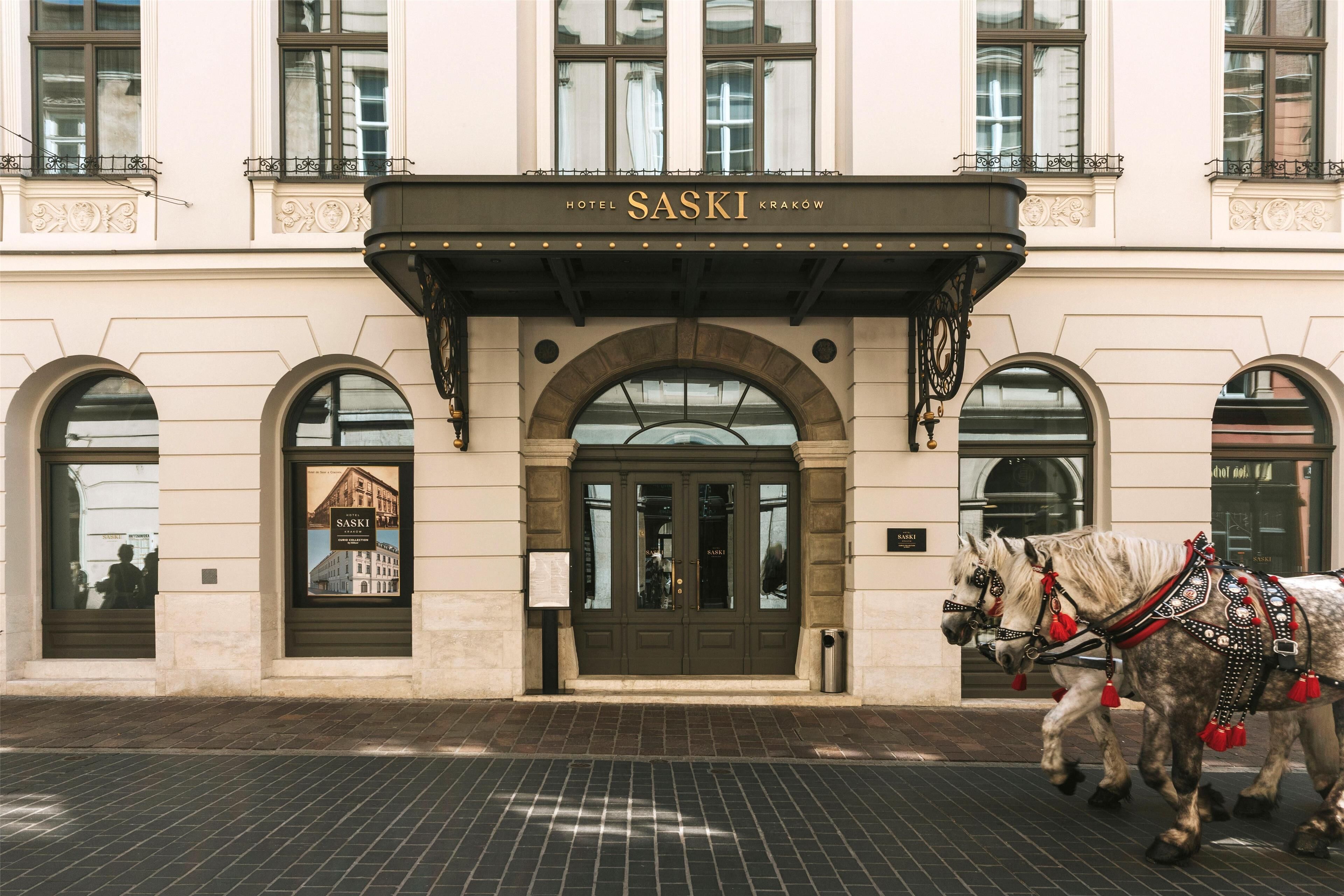 A building with a sign reading 'HOTEL SASKI KRAKÓW' above the entrance. There are two horses with decorative harnesses in front of the building.