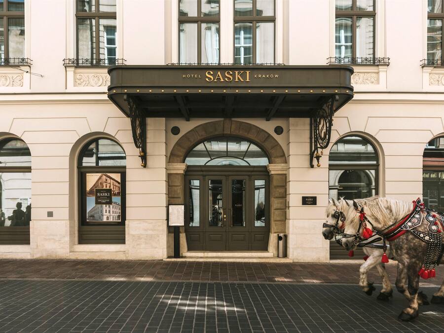 Un edificio con un letrero que dice 'HOTEL SASKI KRAKÓW' sobre la entrada. Hay dos caballos con arneses decorativos frente al edificio.