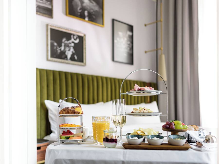 Ontbijt met een etagère, een glas sap en diverse soorten fruit en gebak op een tafel in een hotelkamer.