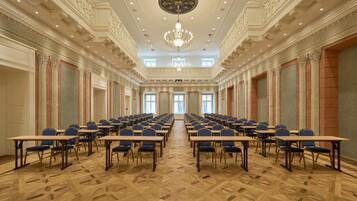 A conference room with rows of tables and chairs, ornate ceiling details, and a chandelier.