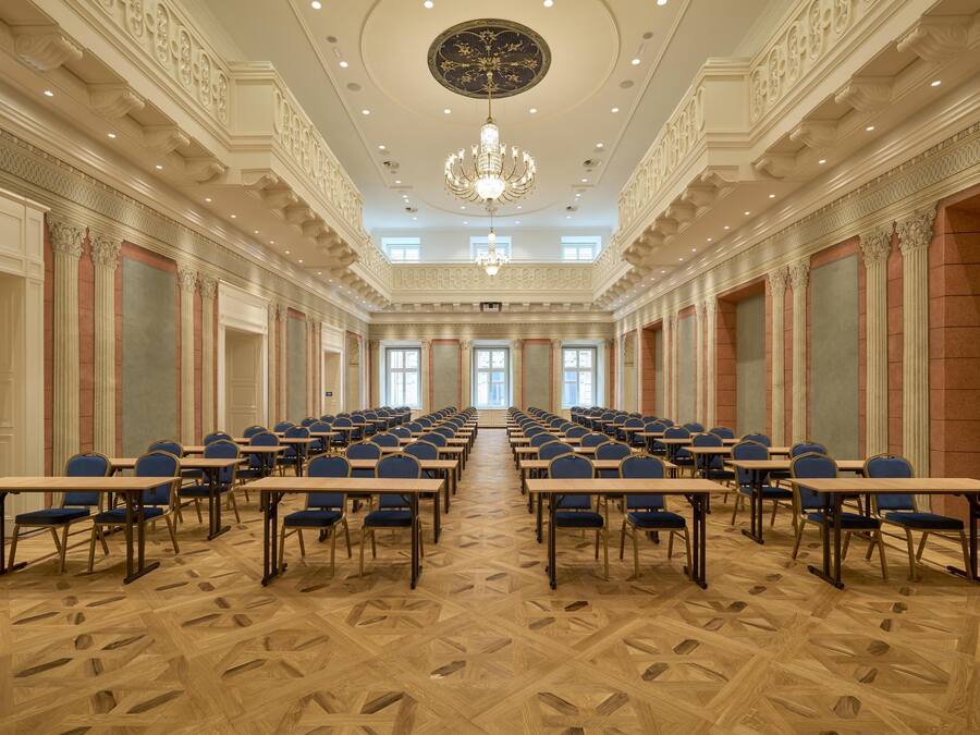 A conference room with rows of tables and chairs, ornate ceiling details, and a chandelier.