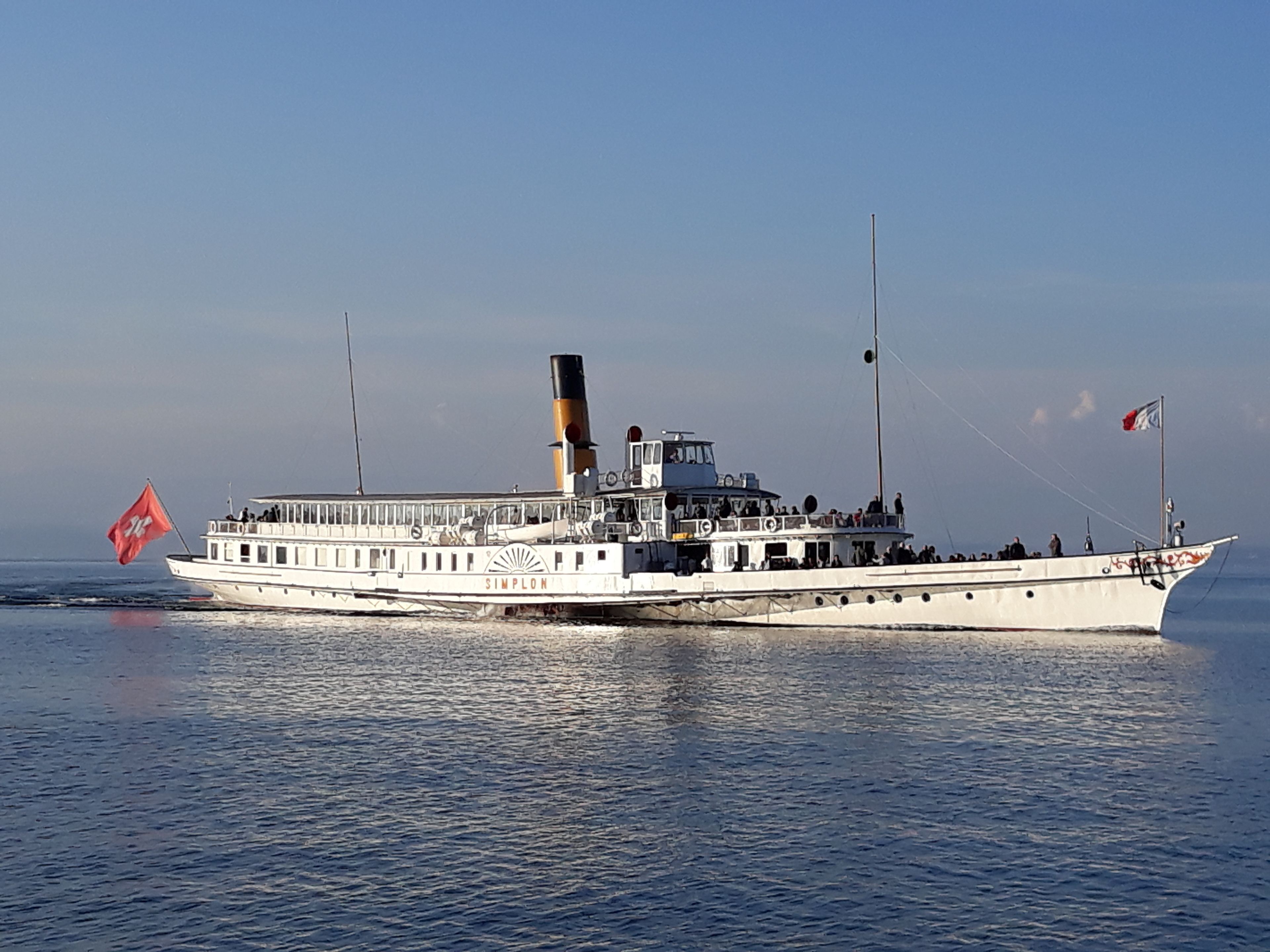 Un grand bateau à vapeur blanc, doté de plusieurs ponts et d’une cheminée imposante, naviguant sur des eaux calmes.