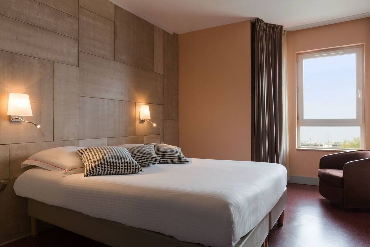 A hotel room with a large bed, two wall-mounted lamps, a window with curtains, and a chair.