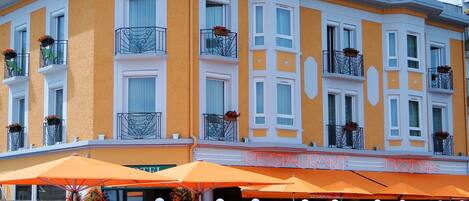 A hotel with a yellow facade, balconies with flower boxes, and outdoor seating with orange umbrellas.