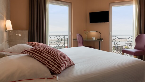 A hotel room with a large bed, two windows with curtains, a flat-screen TV, a small table with a lamp, and a chair.